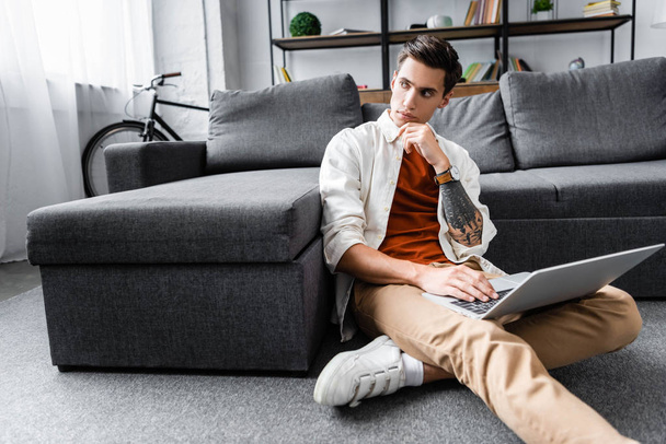 handsome man in shirt sitting on floor and using laptop in apartment  - Foto, Imagen