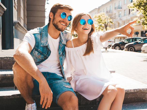 Smiling beautiful girl and her handsome boyfriend.Woman in casual summer dress and man in jeans clothes. Happy cheerful family.Sitting on stairs in street.Points on something interesting.In sunglasses - Photo, Image