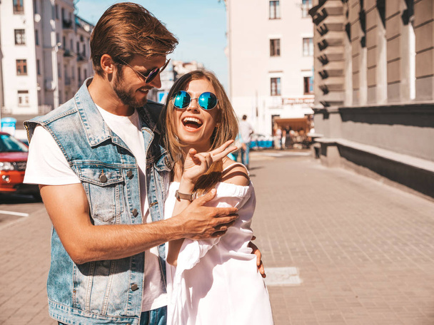 Smiling beautiful girl and her handsome boyfriend walking in the street. Woman in casual summer dress and man in jeans clothes. Happy cheerful couple family having fun in sunglasses - Photo, image