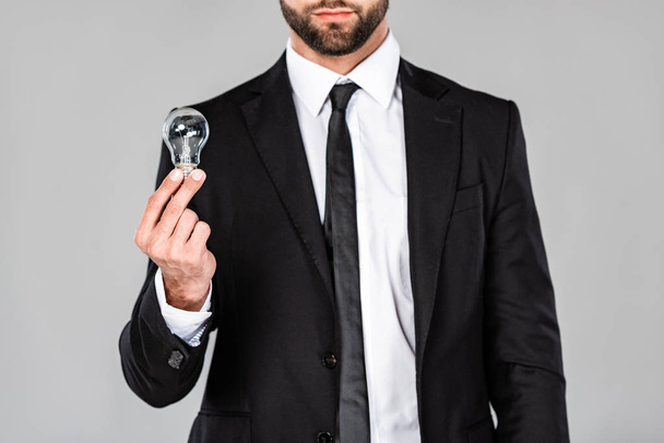 vista parcial del exitoso hombre de negocios en traje negro sosteniendo bombilla aislada en gris
 - Foto, Imagen