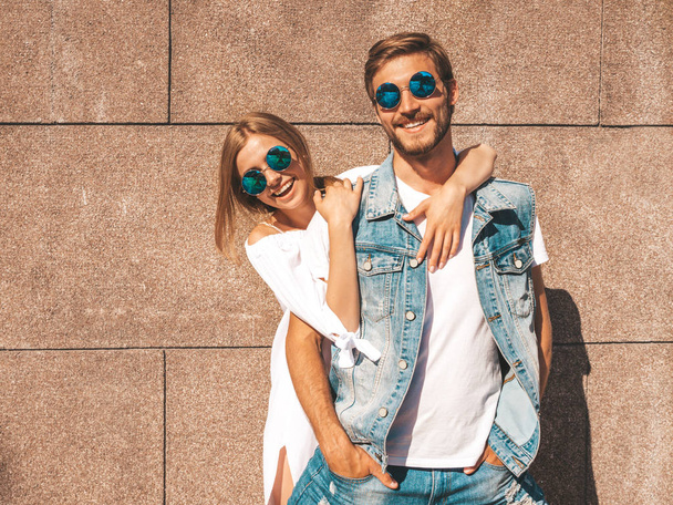 Smiling beautiful girl and her handsome boyfriend. Woman in casual summer dress.Man in jeans clothes.Happy cheerful family.Female having fun on the street near wall.Hugging couple in sunglasses - Zdjęcie, obraz