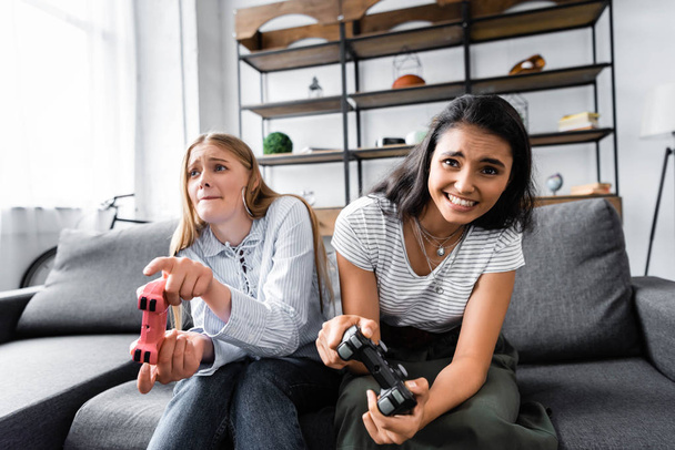 KYIV, UCRANIA - 10 de julio de 2019: amigos multiculturales sentados en el sofá y jugando videojuegos en el apartamento
  - Foto, imagen