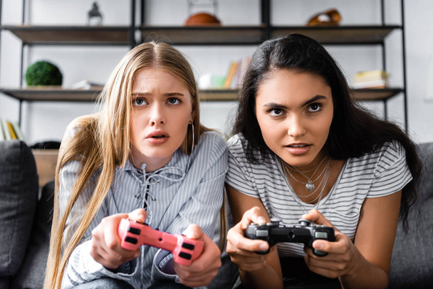 KYIV, UKRAINE - JULY 10, 2019: multicultural friends sitting on sofa and playing video game in apartment  - Foto, Imagen