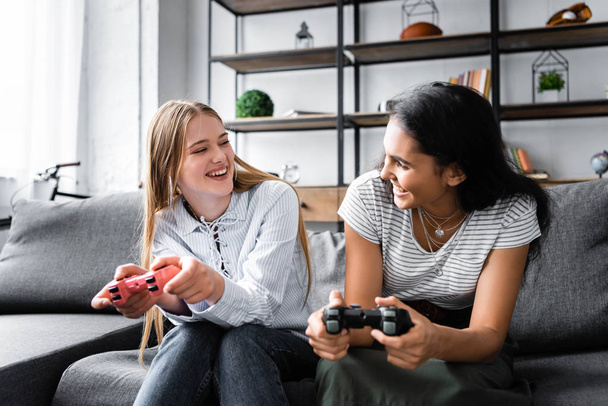 KYIV, UKRAINE - JULY 10, 2019: multicultural friends sitting on sofa and playing video game in apartment  - Fotografie, Obrázek
