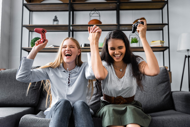 KYIV, UKRAINE - JULY 10, 2019: multicultural friends sitting on sofa and playing video game in apartment  - Foto, Bild
