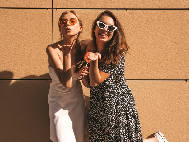 Two young beautiful smiling hipster girls in trendy summer dress. Sexy carefree women posing on street near yellow wall.Positive models having fun in sunglasses.Eating candy lollipop - Foto, Imagem