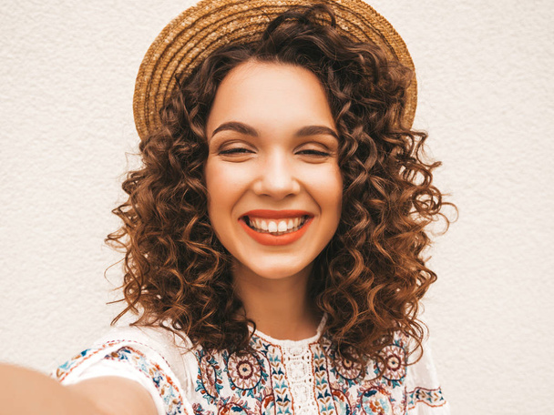 Beautiful smiling model with afro curls hairstyle dressed in summer hipster white dress.Sexy carefree girl posing in the street.Taking selfie self portrait photos in hat on smartphone - Photo, Image