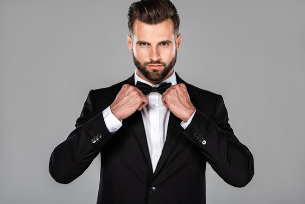 elegant man in black suit fixing bow tie isolated on grey - Фото, изображение