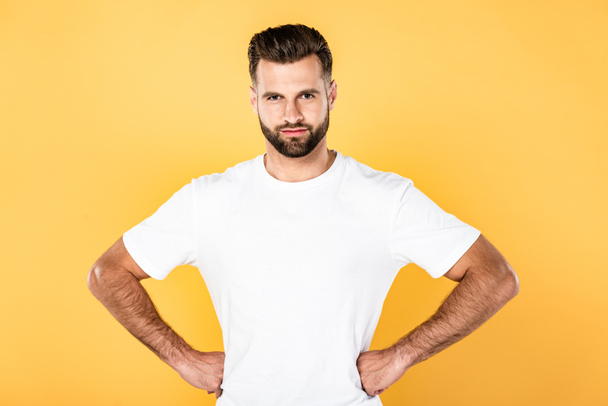 smiling handsome man in white t-shirt with hands on hips isolated on yellow - Foto, afbeelding