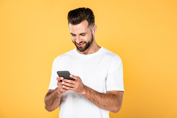 hombre guapo sonriente en camiseta blanca usando teléfono inteligente aislado en amarillo
 - Foto, imagen