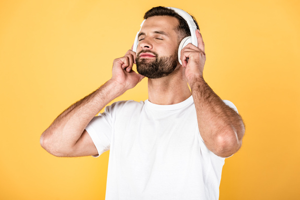 vrolijke man in wit t-shirt luisteren muziek in koptelefoon met gesloten ogen geïsoleerd op geel - Foto, afbeelding