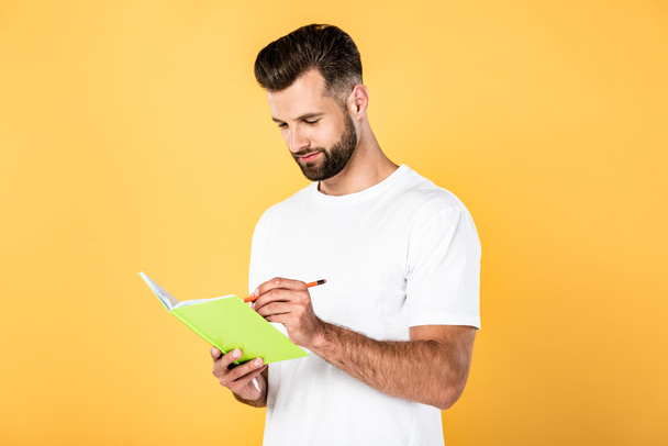 handsome man in white t-shirt writing in notebook isolated on yellow - Photo, Image