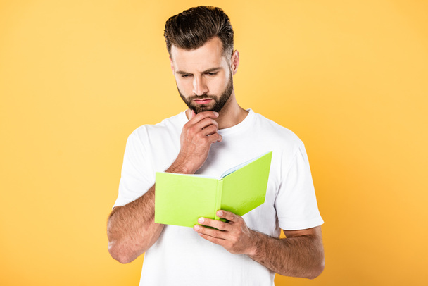 вдумчивый красивый мужчина в белой футболке читает книгу, изолированную на желтый
 - Фото, изображение