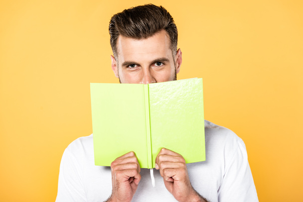 handsome man in white t-shirt holding book in front of face isolated on yellow - Foto, imagen