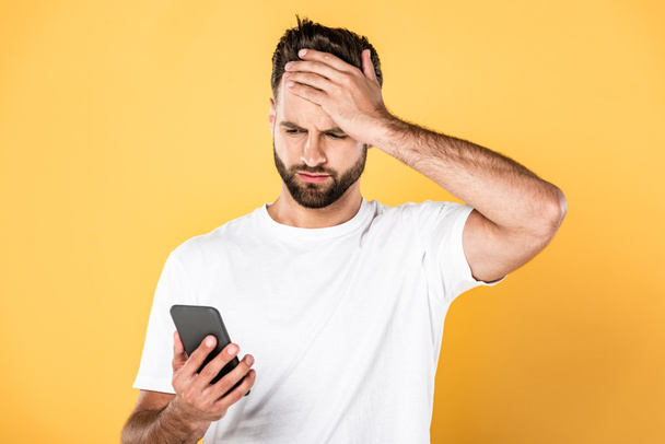 upset man in white t-shirt with facepalm using smartphone isolated on yellow - Foto, Imagen