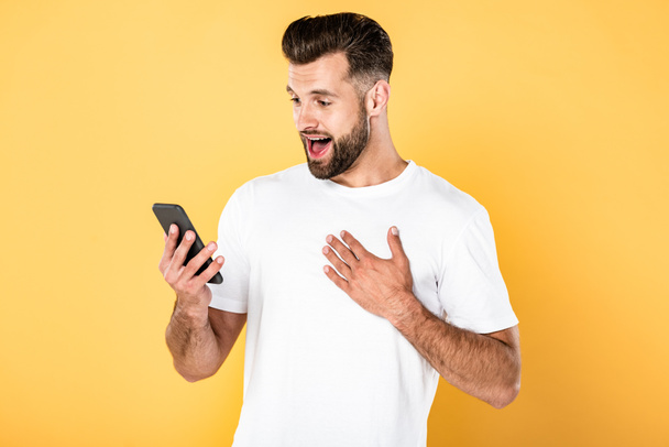 excited handsome man in white t-shirt using smartphone isolated on yellow - Fotoğraf, Görsel