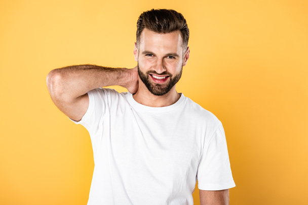 happy handsome man in white t-shirt touching neck isolated on yellow - Фото, изображение
