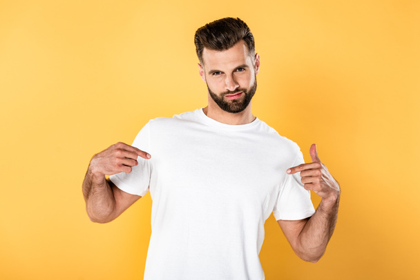 эгоистичный красивый мужчина в белой футболке, указывающий пальцами на себя, изолированный на желтом
 - Фото, изображение
