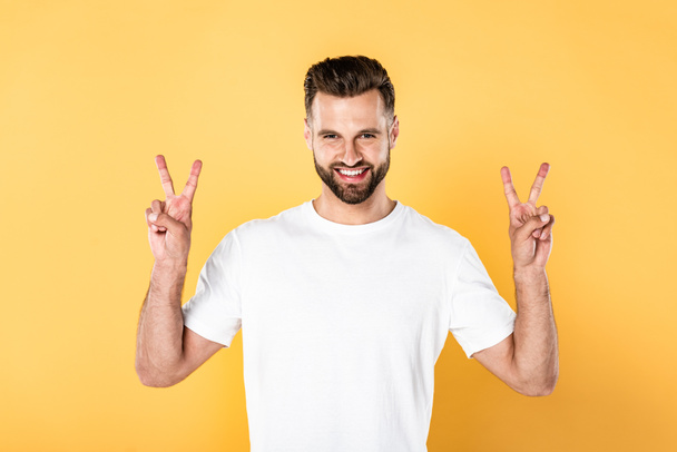 happy handsome man in white t-shirt looking at camera and showing peace signs isolated on yellow - Photo, Image