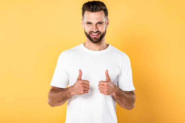 hombre guapo sonriente en camiseta blanca mirando a la cámara y mostrando los pulgares hacia arriba aislado en amarillo
 - Foto, imagen