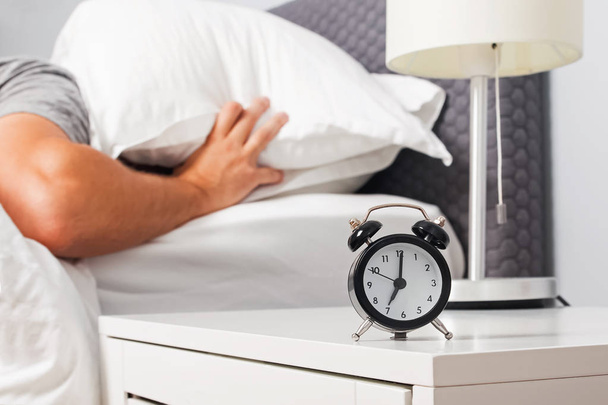 Alarm clock on the nightstand and man closing his had with pillow on the background. - Фото, изображение