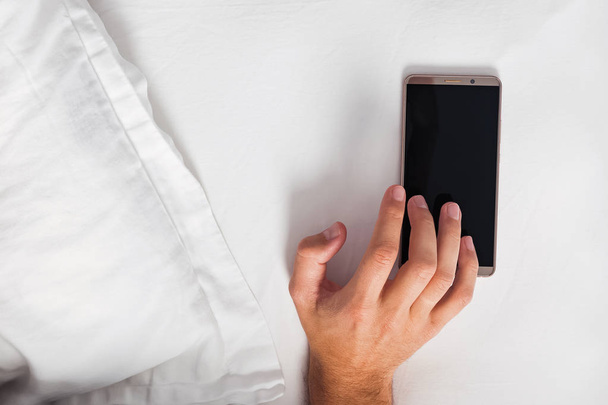 Hand touching the smartphone lying in the bed near the pillow. - Foto, immagini