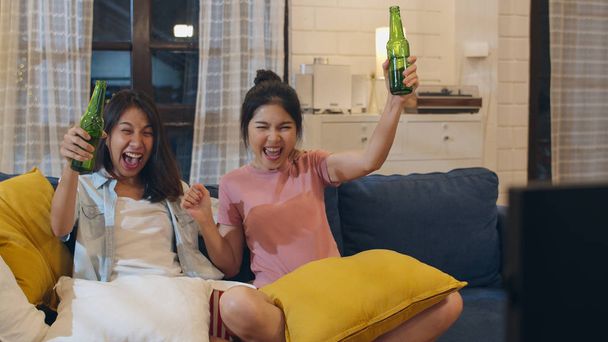Lesbianas lgbt mujeres pareja fiesta en casa, Asiática mujer bebiendo cerveza viendo TV animar fútbol divertido momento juntos en sofá en sala de estar por la noche. Joven amante del fútbol, celebrar el concepto de vacaciones
. - Foto, Imagen