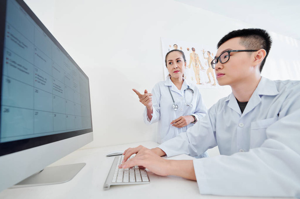 Médico vietnamita e seu assistente discutindo calendário com clientes compromissos na tela do computador
 - Foto, Imagem