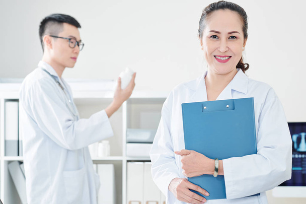 Cheerful mature Vietnamese doctor with clipboard smiling and looking at camera - Photo, image