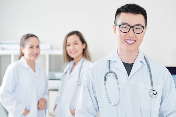Szczęśliwy młody wietnamski lekarza w okularach uśmiechnięty w aparacie, jego koledzy stojący w tle - Zdjęcie, obraz