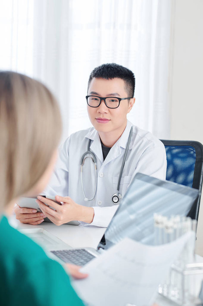 Young Vietnamese doctor in glasses sitting at table with digital tablet in hands and talking to physician - Fotografie, Obrázek