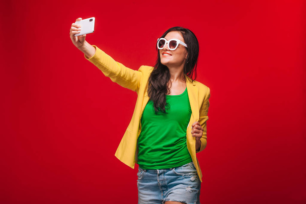 Κορίτσι σε ένα κόκκινο φόντο στο μπουφάν παίρνει μια selfie στο τηλέφωνο - Φωτογραφία, εικόνα