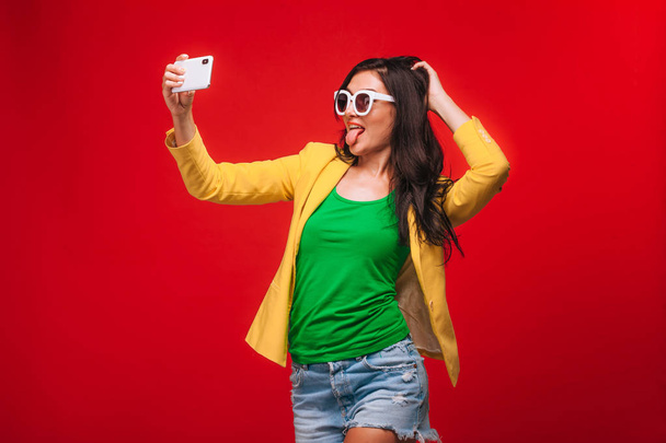 Mädchen auf rotem Hintergrund in Jacke macht ein Selfie mit dem Handy - Foto, Bild