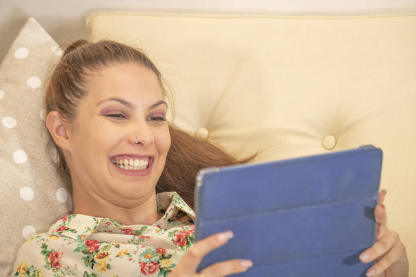 Retrato de una mujer blanca sonriente con el pelo castaño, alegre viendo videos divertidos en su tableta azul
. - Foto, Imagen