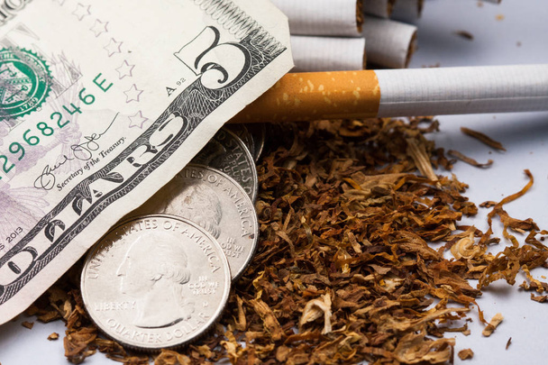 Cigarettes separated with some shredded tobacco scattered, with pennies around and some dollars, isolated on black background. Tobacco can cause diseases. - Photo, Image