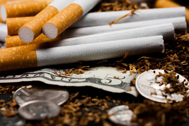 Savukkeet erotettu silputtu tupakka hajallaan, pennejä ympärillä ja dollareita, eristetty mustalla pohjalla. Tupakka voi aiheuttaa sairauksia
. - Valokuva, kuva