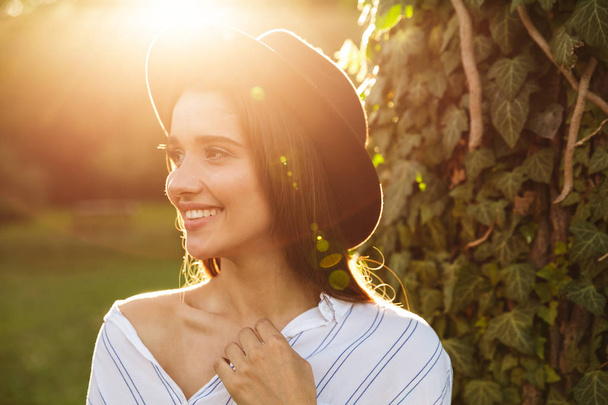 Foto de uma jovem mulher bonita usando chapéu elegante sorrindo e andando no parque verde
 - Foto, Imagem