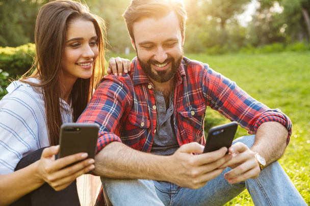 Image of attractive couple man and woman using cellphones while resting in green park - Zdjęcie, obraz