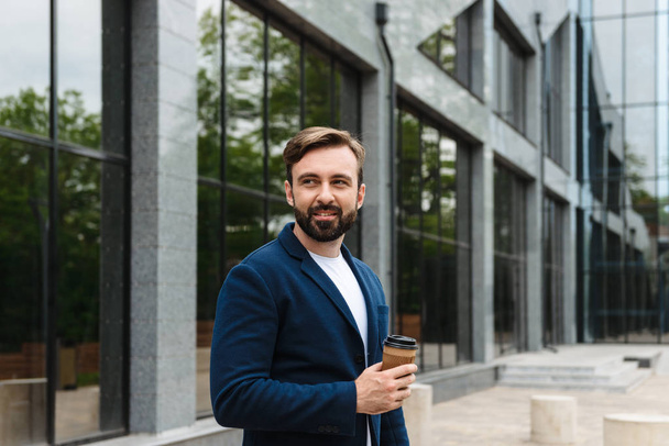 Portrait of brunette businessman in jacket holding takeaway paper cup with coffee while standing outdoors - Фото, изображение