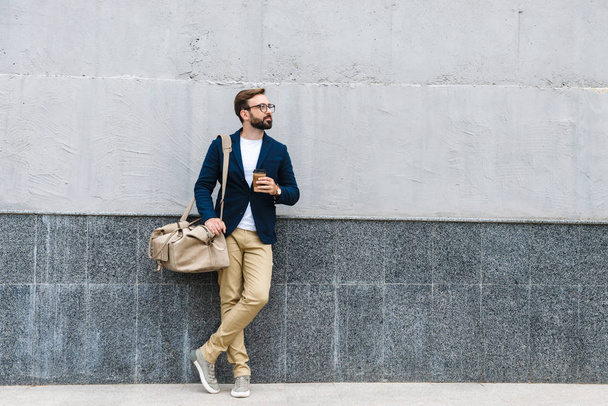 Portrait of unshaven businessman wearing eyeglasses drinking coffee from paper cup and carrying bag while standing near wall - Fotografie, Obrázek