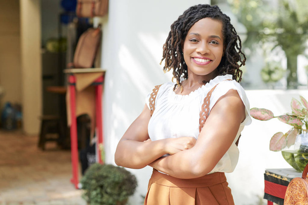 Retrato de una hermosa mujer africana de pie con los brazos cruzados y sonriendo a la cámara cerca de la tienda al aire libre
 - Foto, imagen