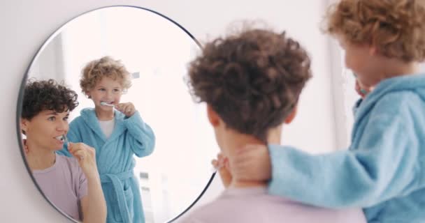Mujer joven y niño pequeño cepillarse los dientes en el baño mirando el espejo
 - Metraje, vídeo