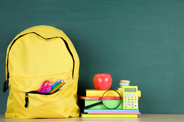 School supplies with yellow backpack on chalkboard background - Foto, afbeelding