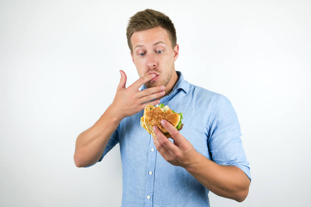 Молодой красивый мужчина наслаждается чизбургером из ресторана быстрого питания, облизывая пальцы, пока ест на изолированном белом фоне.
 - Фото, изображение