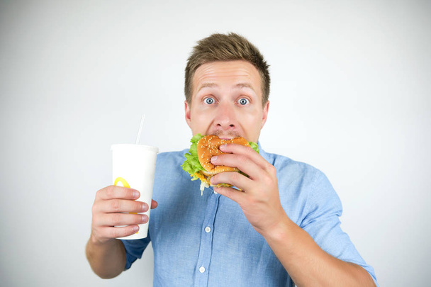 Молодой красивый мужчина кусает чизбургер из ресторана быстрого питания и держит газировку чувство голода на изолированном белом фоне
 - Фото, изображение