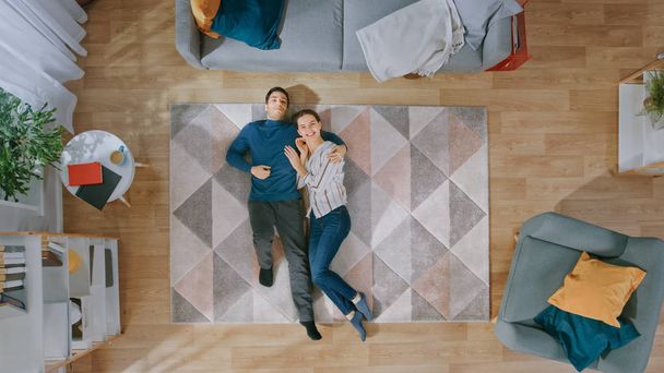 Young Happy Couple is Lying Down on the Floor and Laughing. Man Hugs the Girl. Cozy Living Room with Modern Interior with Carpet, Sofa, Chair, Table, Shelf, Plants and Wooden Floor. Top Down. - Фото, зображення