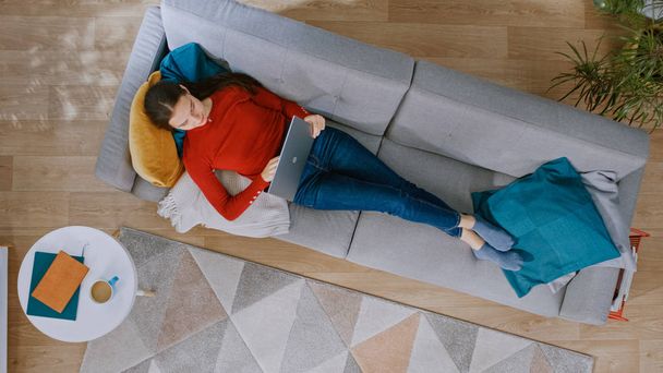 Young Girl in Red Jumper and Blue Jeans is Lying Down on a Couch, Working on a Laptop. Looks Above, Smiles. Cozy Living Room with Modern Interior with Plants, Coffee Table and Wooden Floor. Top View. - Фото, изображение