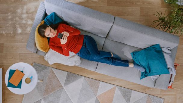 Young Girl in Red Jumper and Blue Jeans is Lying Down on a Sofa, Using a Smartphone. She is Happy and Smiles. Cozy Living Room with Modern Interior with Carpet, Coffee Table and Wooden Floor. Top View - Φωτογραφία, εικόνα