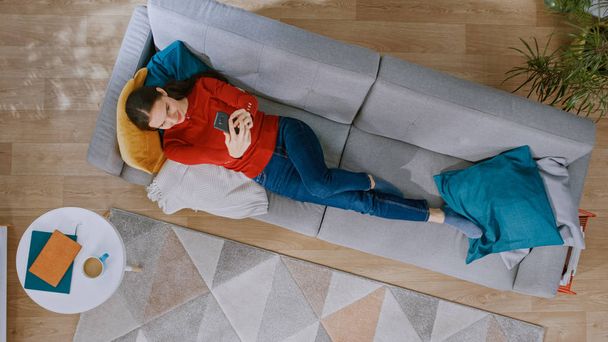 Young Girl in Red Jumper and Blue Jeans is Lying Down on a Sofa, Using a Smartphone. She is Happy and Smiles. Cozy Living Room with Modern Interior with Carpet, Coffee Table and Wooden Floor. Top View - Foto, imagen