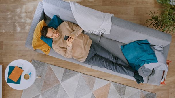 Young Man in Brown Jumper and Grey Jeans is Lying Down on a Sofa, Using a Smartphone. He is Happy and Smiles. Cozy Living Room with Modern Interior with Carpet, Plants, Table, Wooden Floor. Top View. - Photo, Image